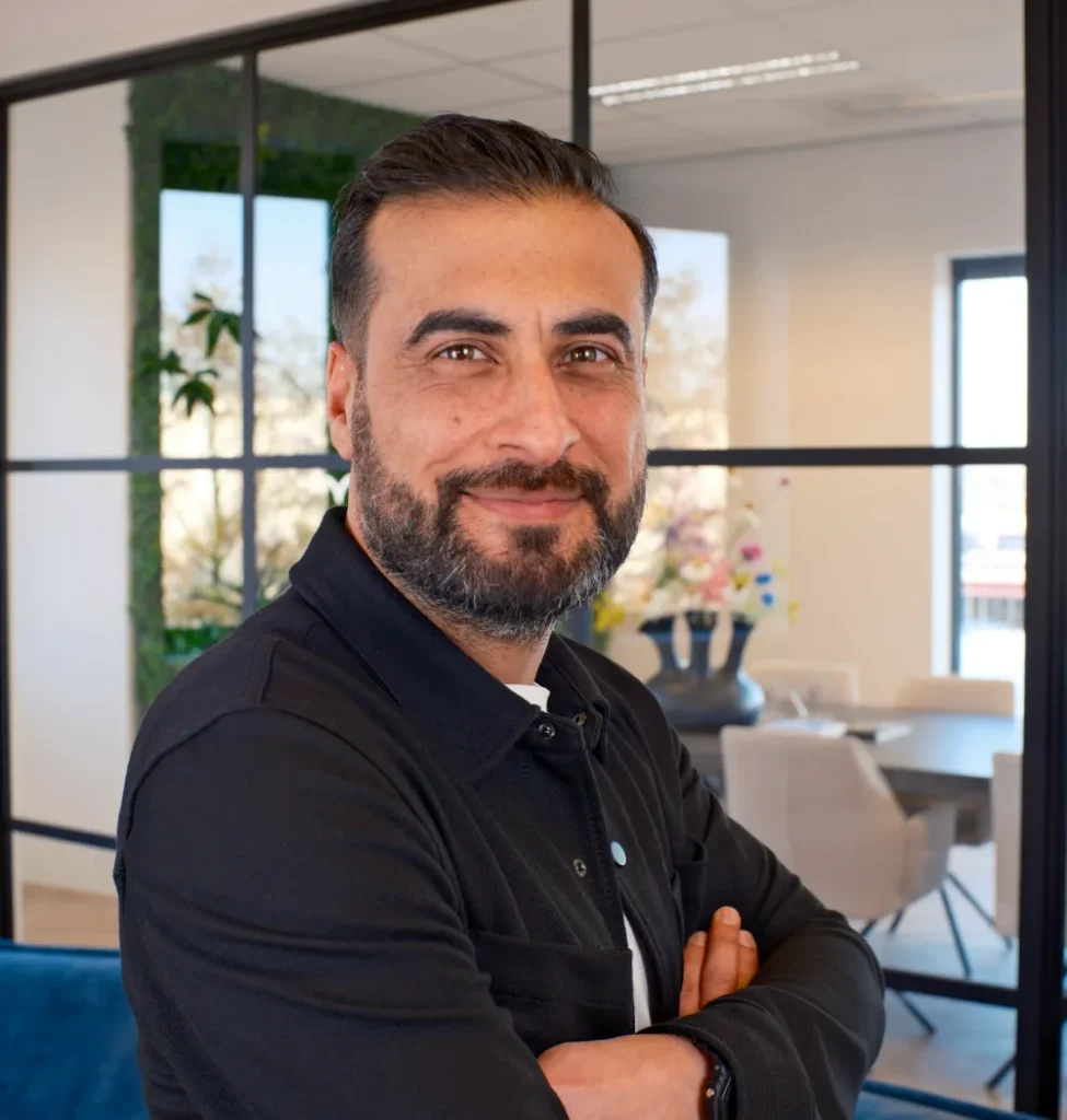 Man met verzorgde baard en zwart overhemd staat met armen gekruist voor glazen wanden en kantoorinrichting