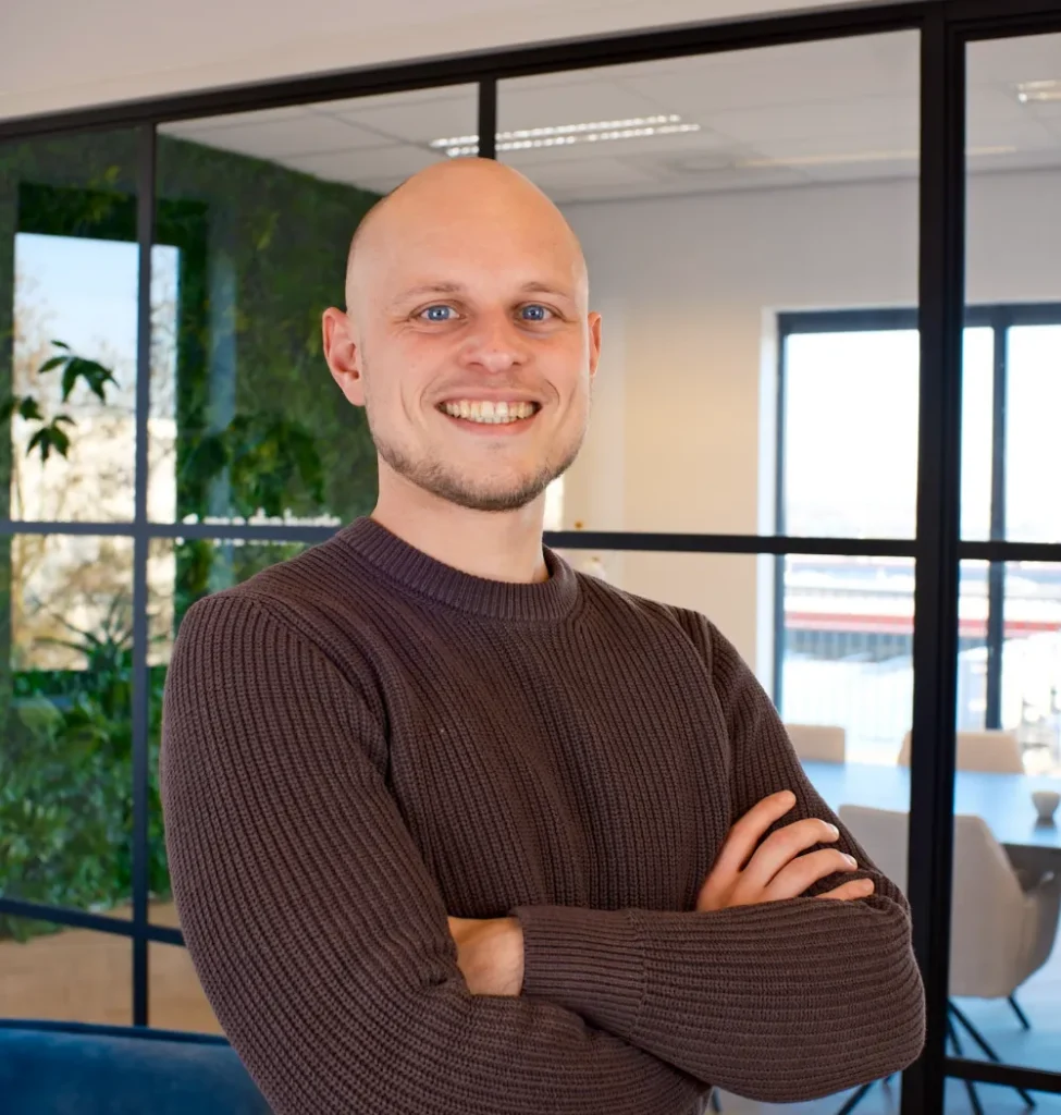 Man met kaal hoofd en glimlach in donkerbruine gebreide trui met kruiselende armen in kantoor