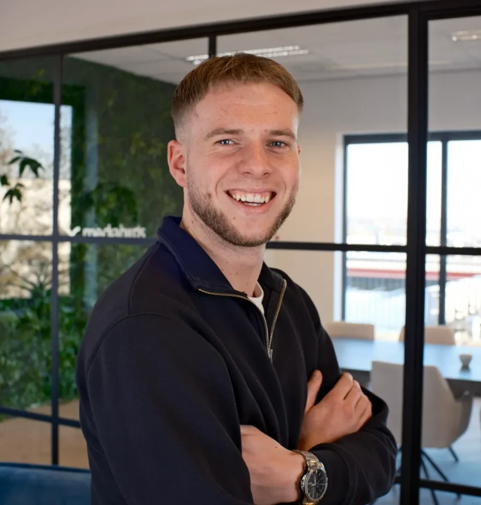 Een jonge man met lichtbruin haar en baard staat met armen gekruist in een modern kantoor.
