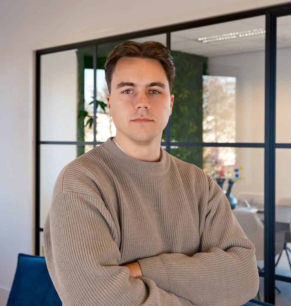 Jongeman in lichtbruine gebreide trui staat met armen over elkaar in modern kantoor met grote ramen.