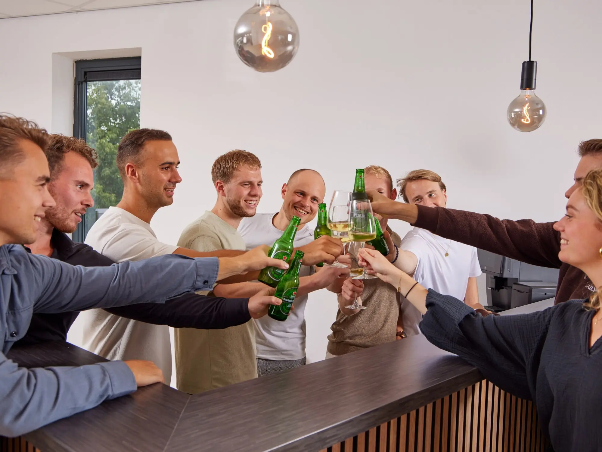 Groep van acht mensen, voornamelijk mannen, proostend met glazen en flessen achter een moderne bar met natuurlijk licht.