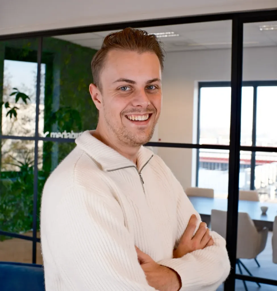 Jonge man met witte trui met hoge kraag staat met gekruiste armen in modern kantoor met grote ramen en groen.