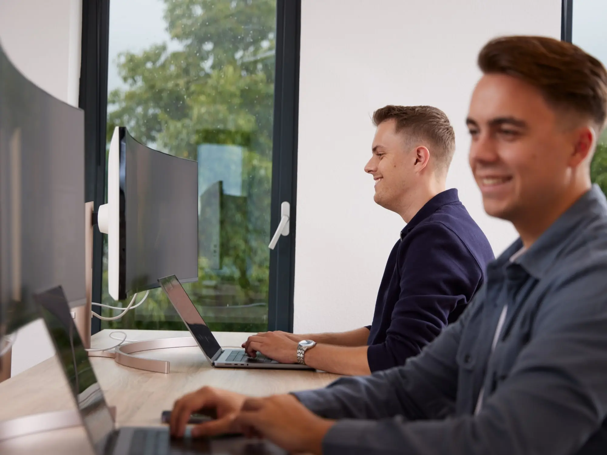Twee jonge mannen die aan een werktafel zitten en geconcentreerd op laptops werken in een lichte kantoorruimte.