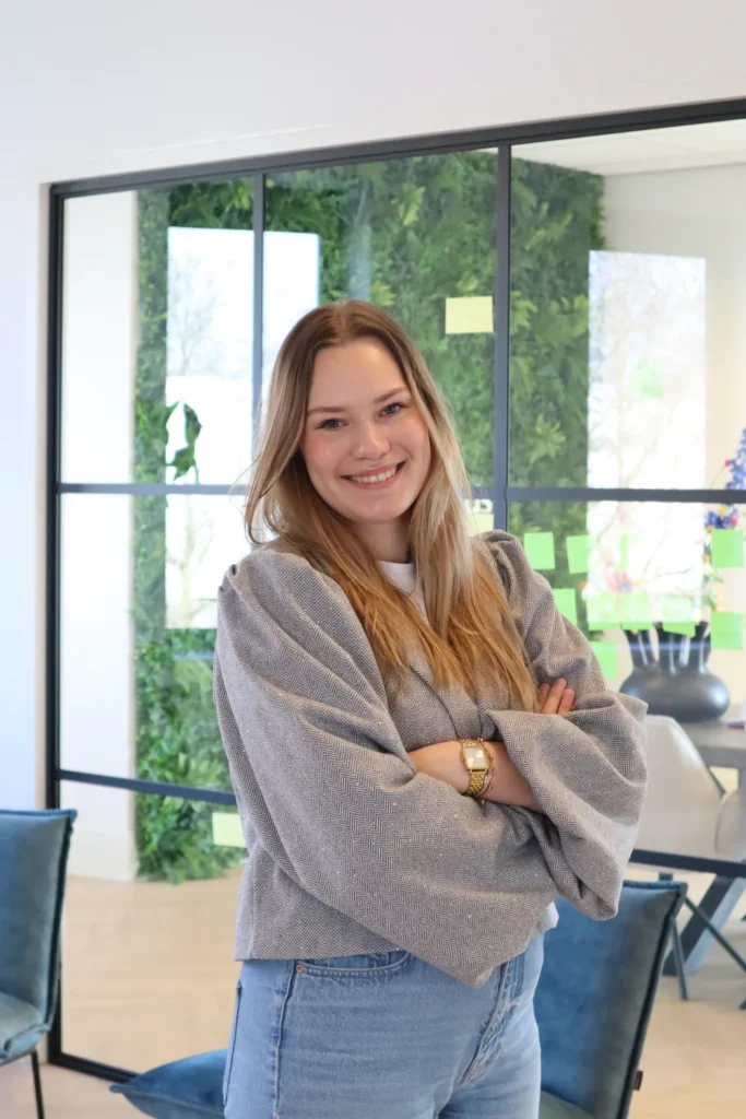 Een jonge vrouw met lange golvende haren staat in een lichte ruimte met grijze trui en spijkerbroek.