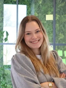 Een jonge vrouw met lange golvende haren staat in een lichte ruimte met grijze trui en spijkerbroek.